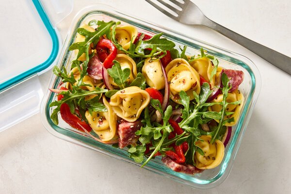 High-Protein Lunch Recipes for Back-to-School An overhead image of a rectangular clear glass container filled with round tortellini pasta, red peppers, salami chunks and arugula leaves.